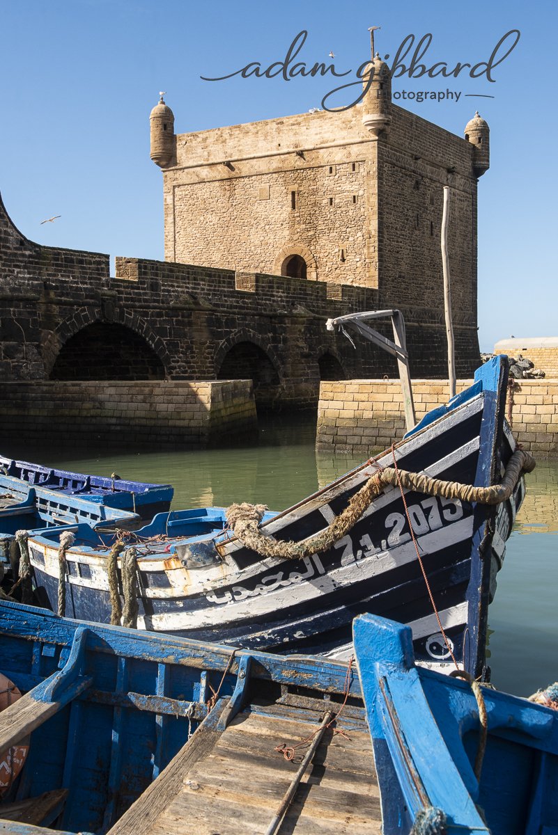 A historic stone fortress with small turrets on its corners stands by a water harbor, with colorful blue boats docked in the foreground.