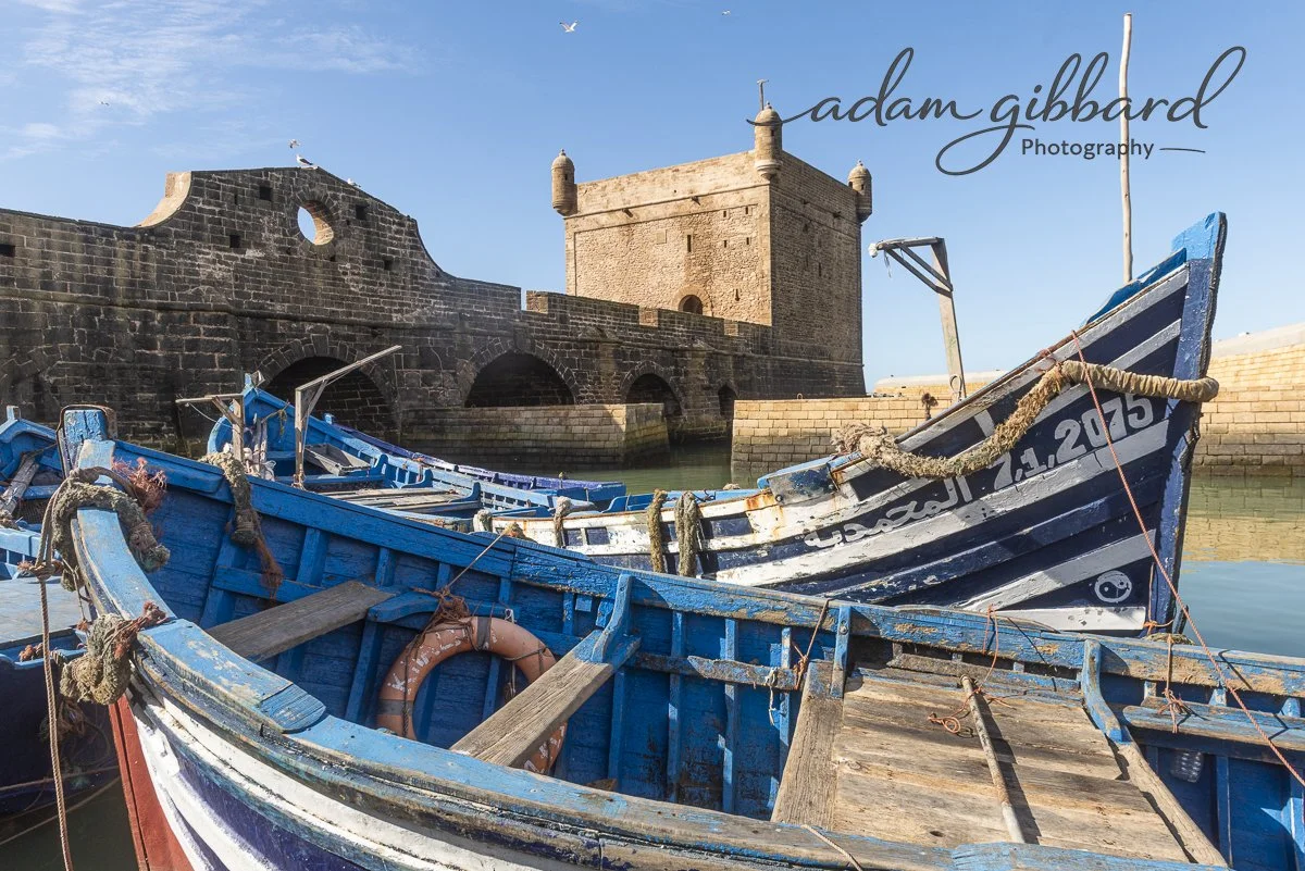 Blue and white boats docked near a stone marina with an old fortress or castle in the background under a clear blue sky.