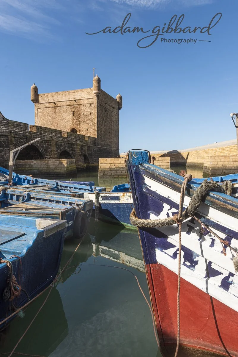 Boats docked near an old stone fortress with a clear blue sky in the background.