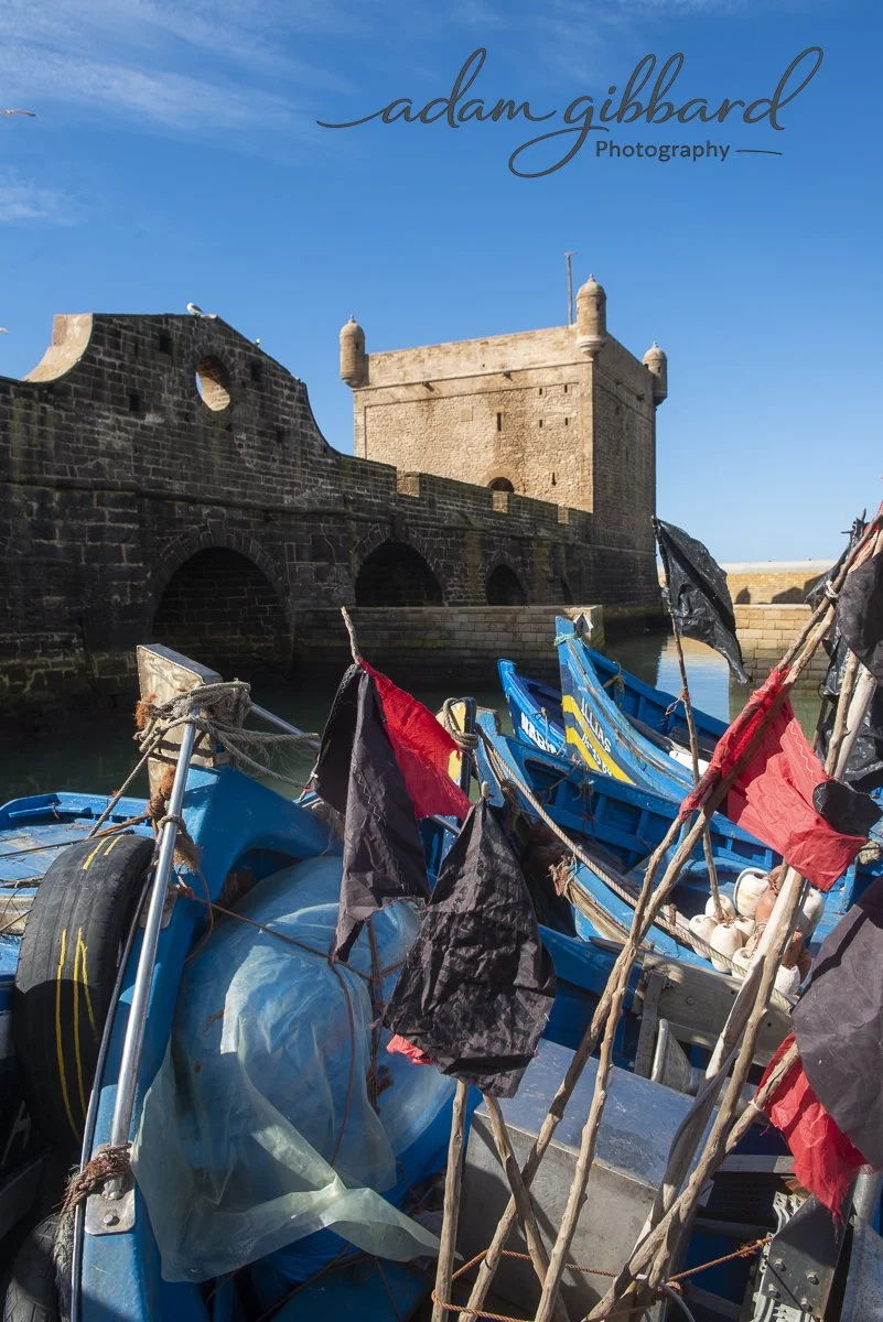 Boats docked near a historic stone fortress under a clear blue sky, with colorful flags and fishing equipment visible in the foreground.
