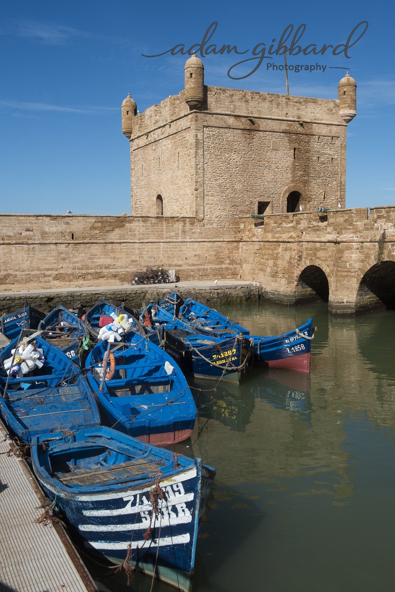 A historic stone fortress with rounded towers sits by the water, with several blue boats moored along the dock in the harbor.