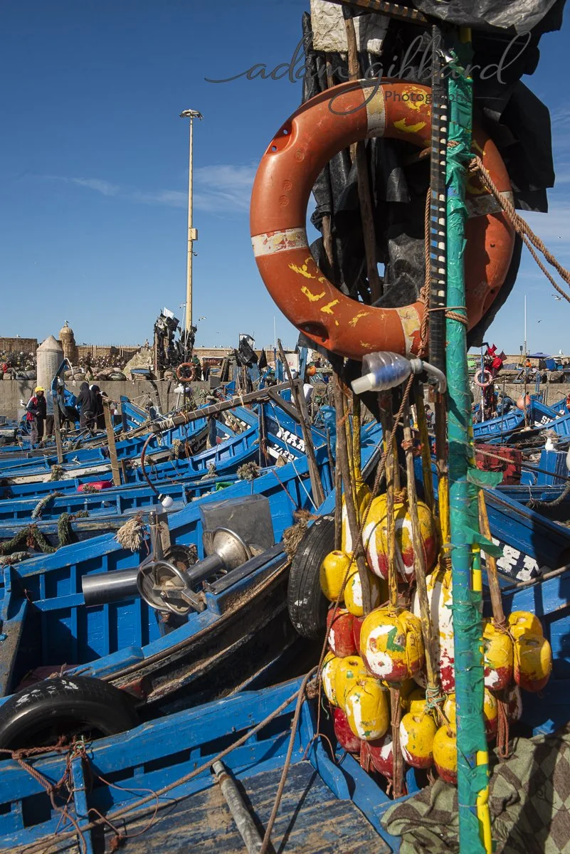 A close-up of a fishing boat dock showing orange rescue ring, yellow and red buoys, and various fishing equipment among blue boats in a busy harbor.