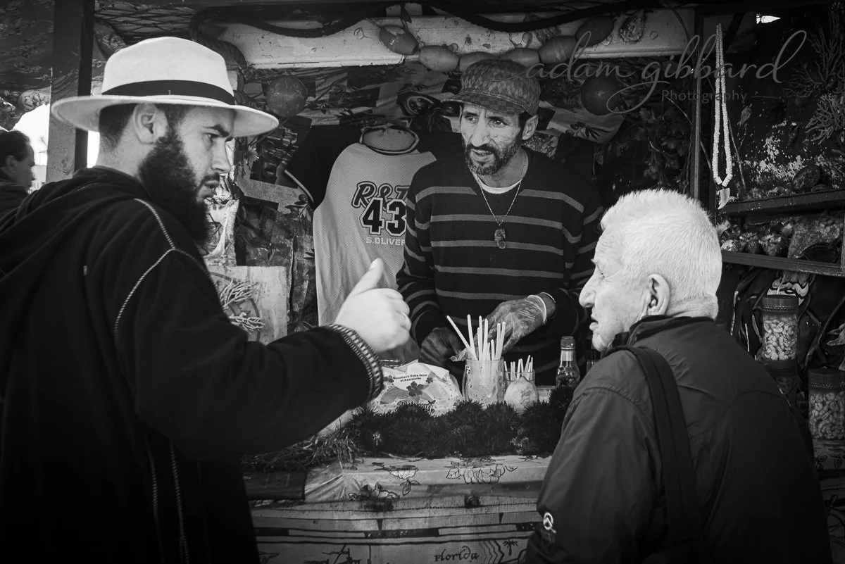 Three men engaged in conversation at a market stall, with food and decorations in the background.