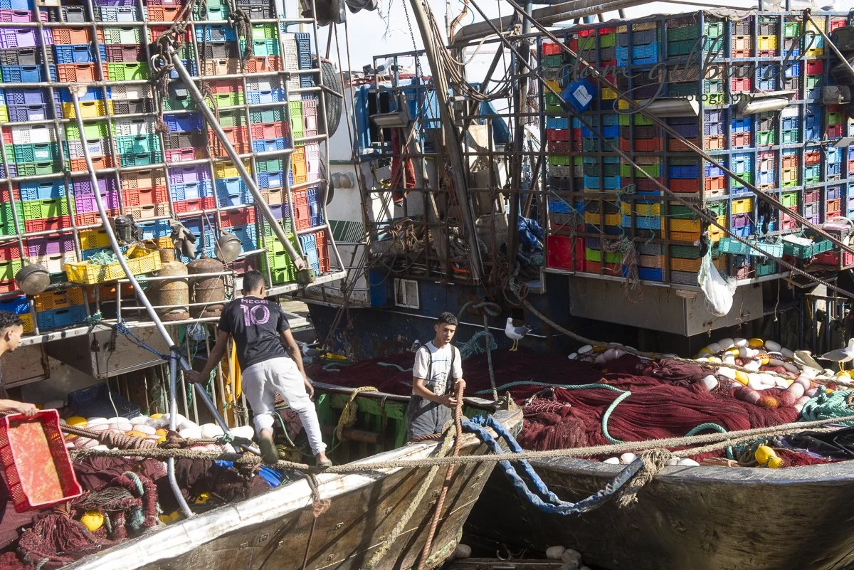 Two boats filled with colorful crates and equipment, with three men working on the boats among ropes and fishing nets, on a dock during daytime.