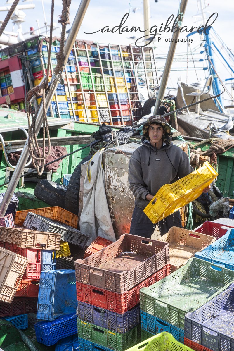 Young man standing among colorful plastic crates and crates stacked on a boat, with more crates and boat equipment visible in the background.