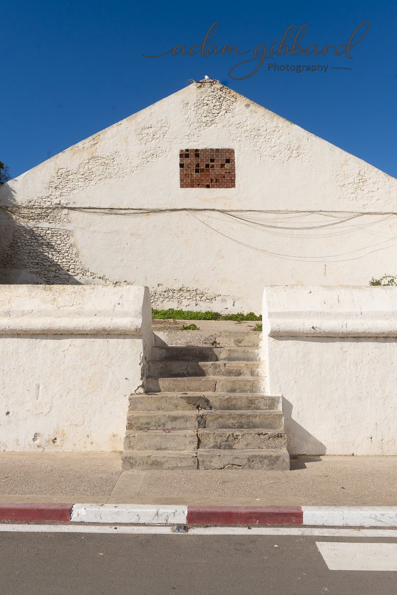 A white stucco building with a triangular roof and a square brick vent in the upper center. There are concrete steps leading up to the building, with a white concrete wall on either side. The sky is clear and blue.