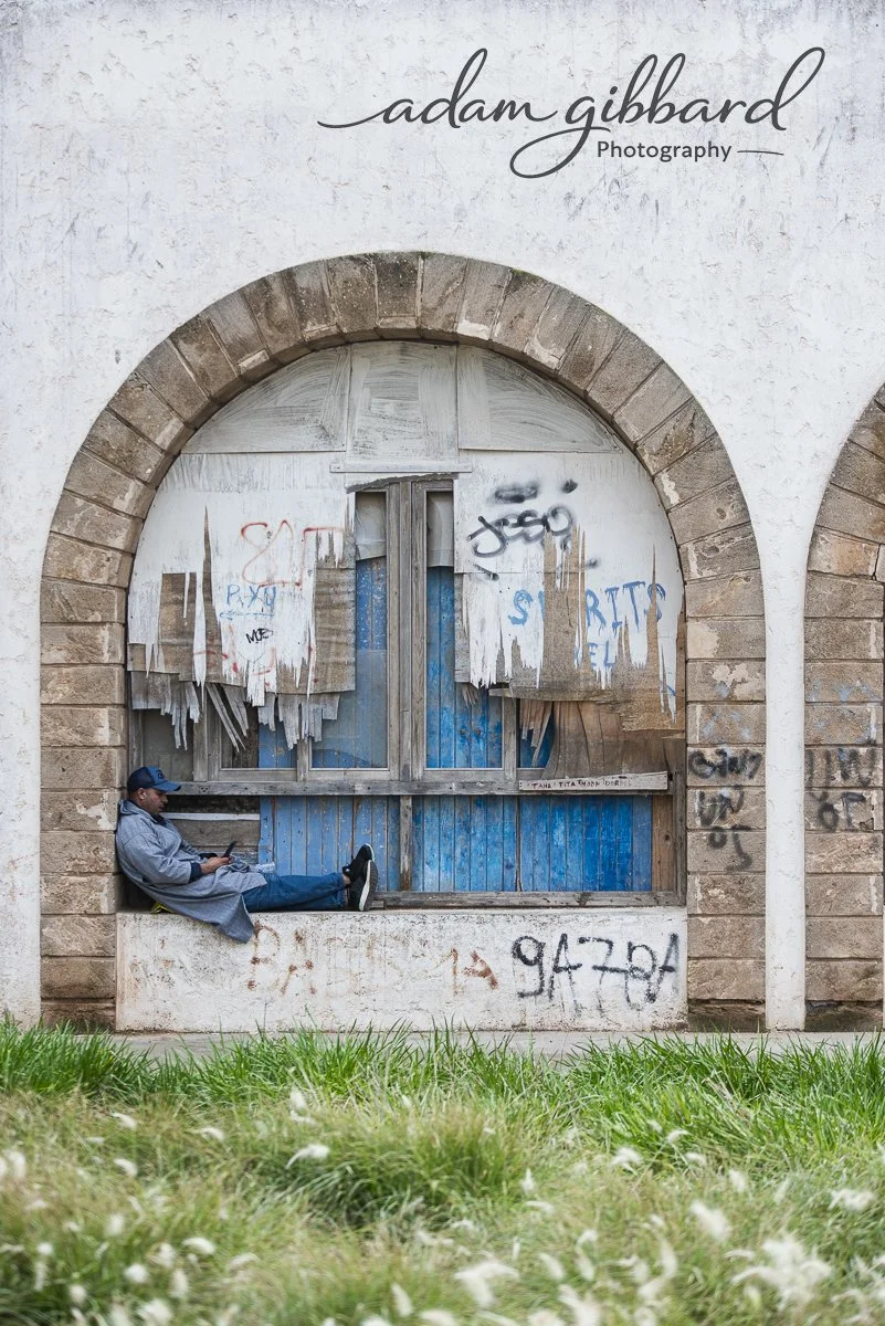 A man sits on a ledge in front of a boarded-up window with peeling paint and graffiti, with grass in the foreground and an arched brick frame around the window.