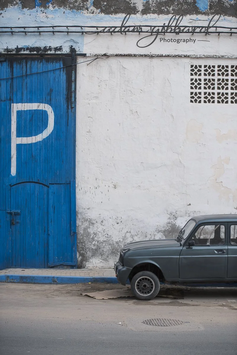 A blue parking garage door with a large white P symbol, a white textured wall with peeling paint, a small lattice window, and a vintage gray car parked on the street.
