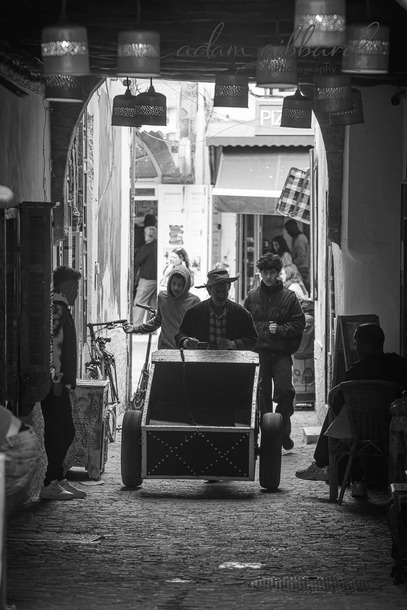 People walking through a narrow alleyway with shops on either side, some sitting and others pushing a cart, black-and-white photo.