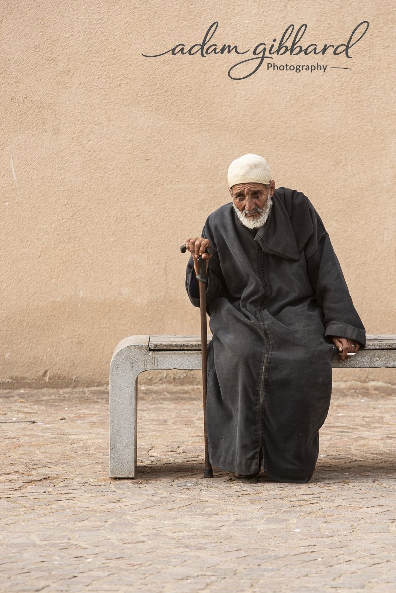 An elderly man with a white beard sitting on a bench against a beige wall, holding a cane, wearing a beige knit cap and a long dark coat.