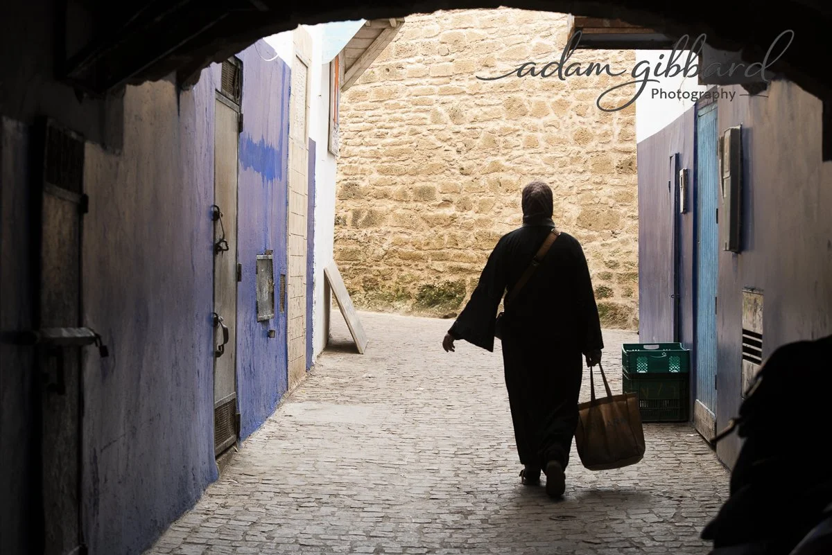 A person wearing a long black outfit and headscarf walking through a narrow alleyway, carrying a brown shopping bag in one hand and a small green crate in the other, seen from behind, with stone and blue-painted walls.