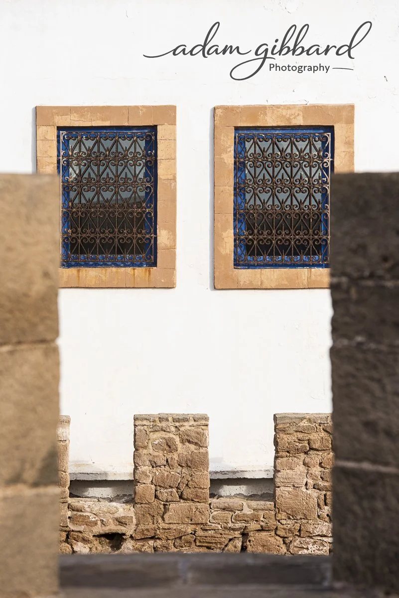 Close-up of two barred windows with brick borders on a white wall, with a sign above reading 'adam gibbard Photography'.