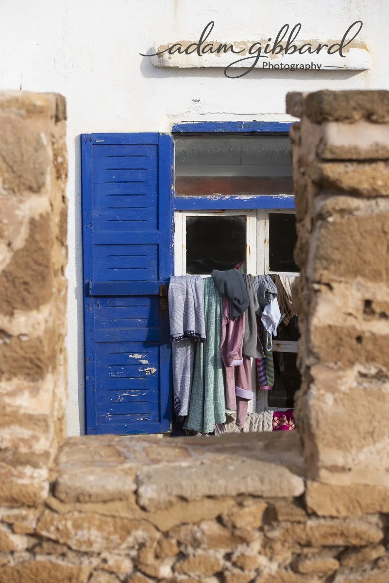 View through a brick opening showing a white building with a blue shutter door and window, and laundry hanging outside.