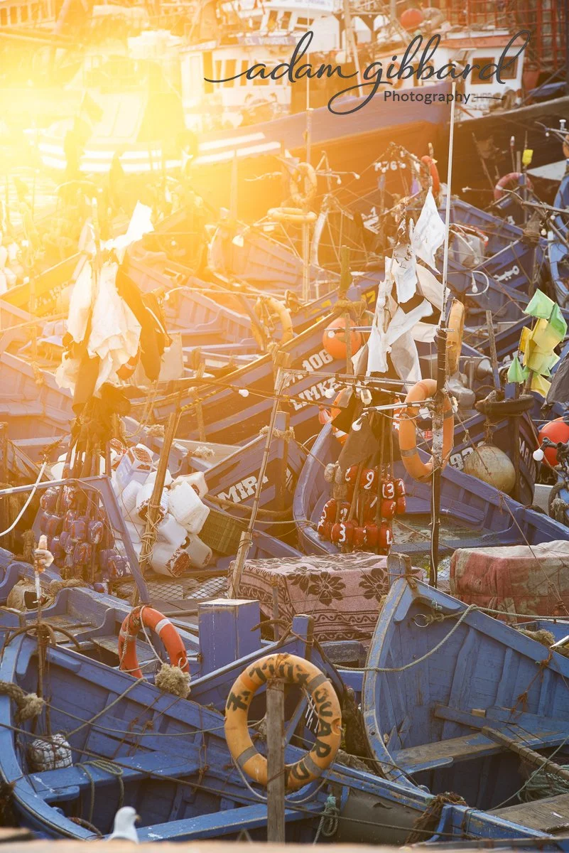 A dock filled with moored boats, with some boats displaying life jackets, ropes, and flags, and a bright sunlight casting a glow over the scene.