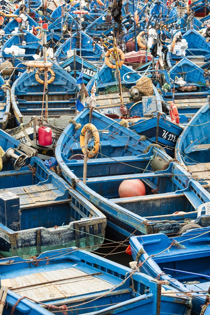 A crowded dock with numerous blue fishing boats tightly packed together, some with fishing gear and floating buoys.
