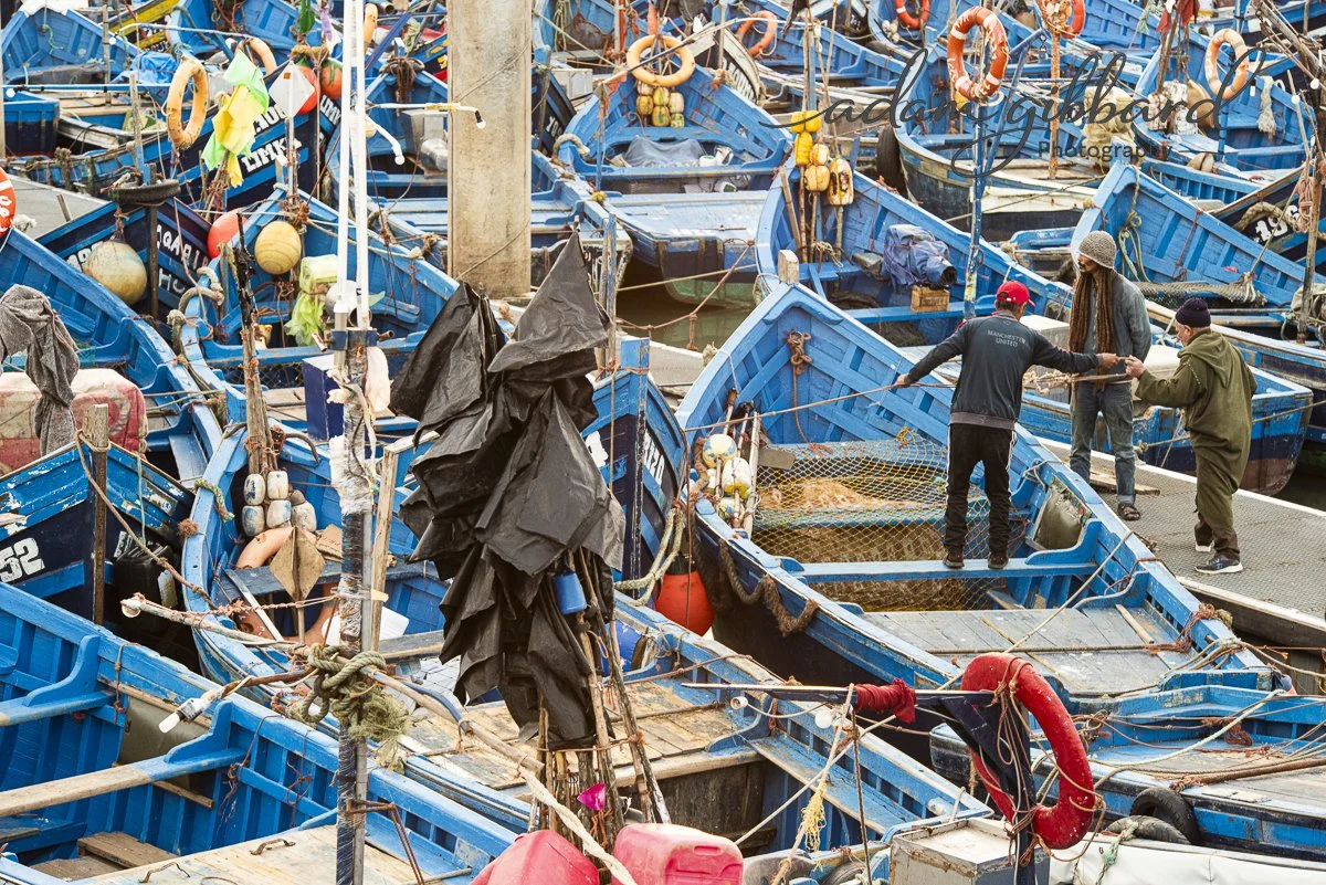 Multiple blue fishing boats docked at a harbor with three men working on the dock, surrounded by fishing gear and equipment.