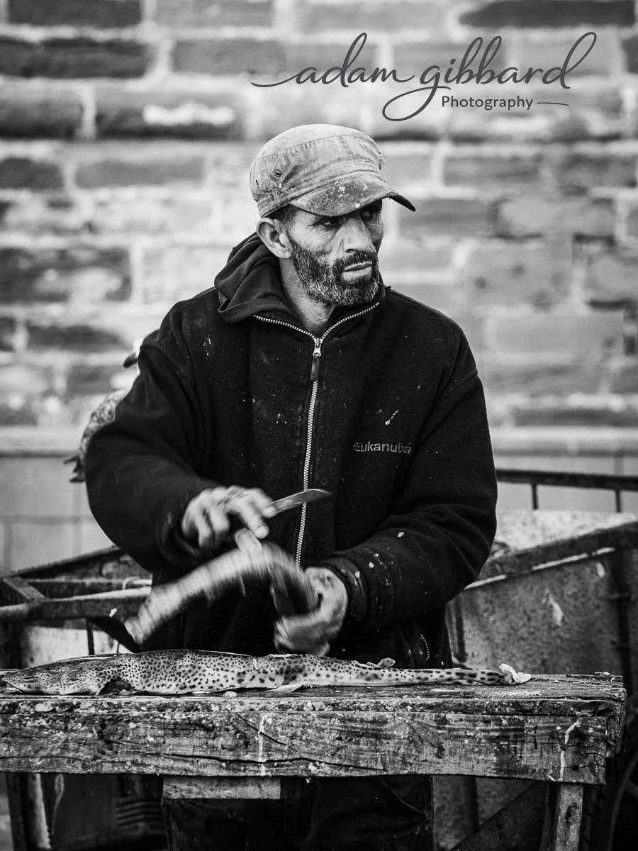 Man dressed in dark hoodie and cap cleaning a fish on a wooden table in a workshop with brick wall background, black and white photograph.