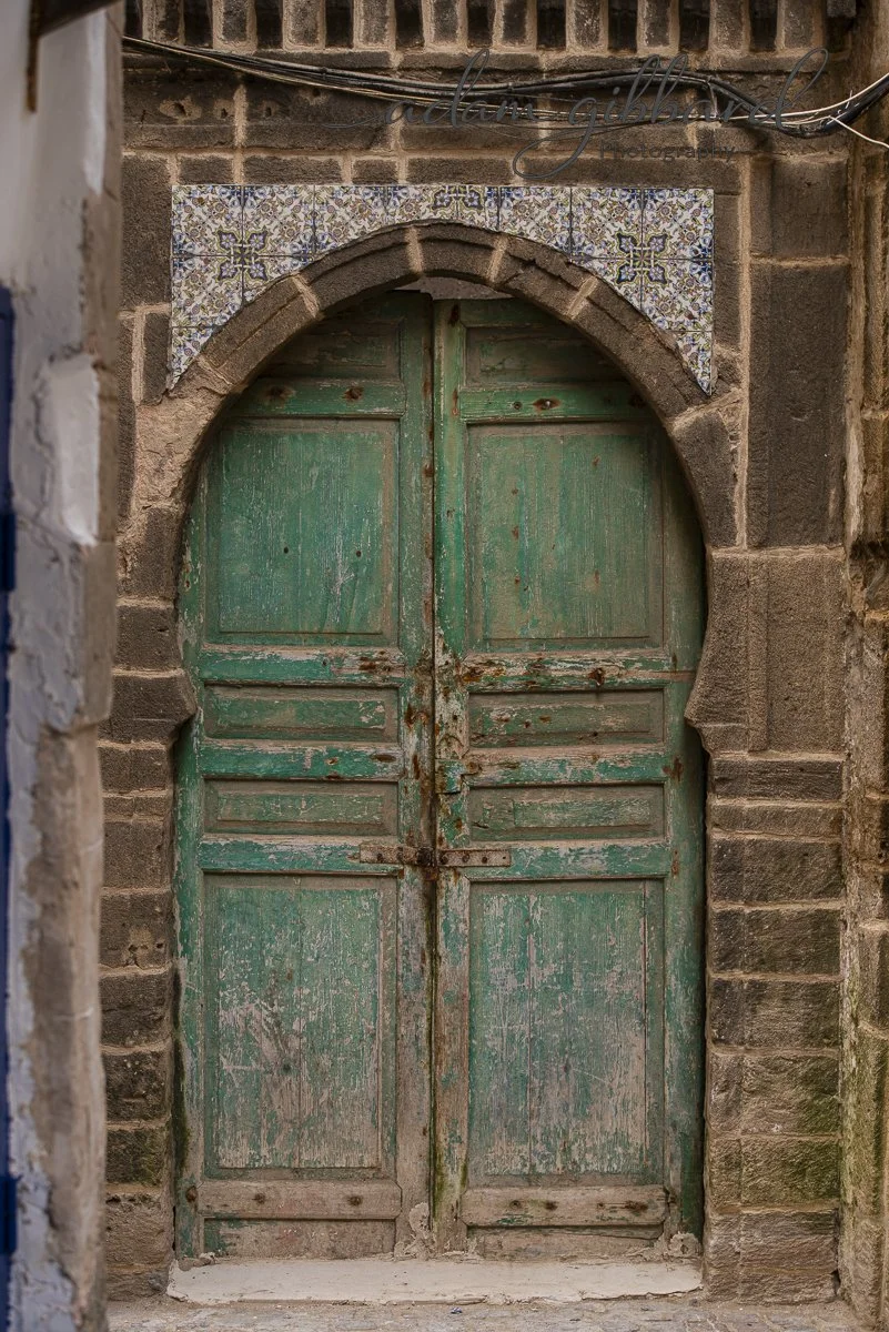 Old, weathered green wooden door with peeling paint set in a brick wall with decorative tile accents above.