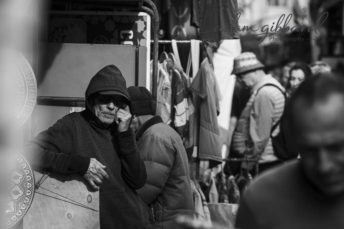 A black and white photo of a man wearing a hoodie and sunglasses, leaning on a countertop or stall at an outdoor market, with other people shopping in the background.