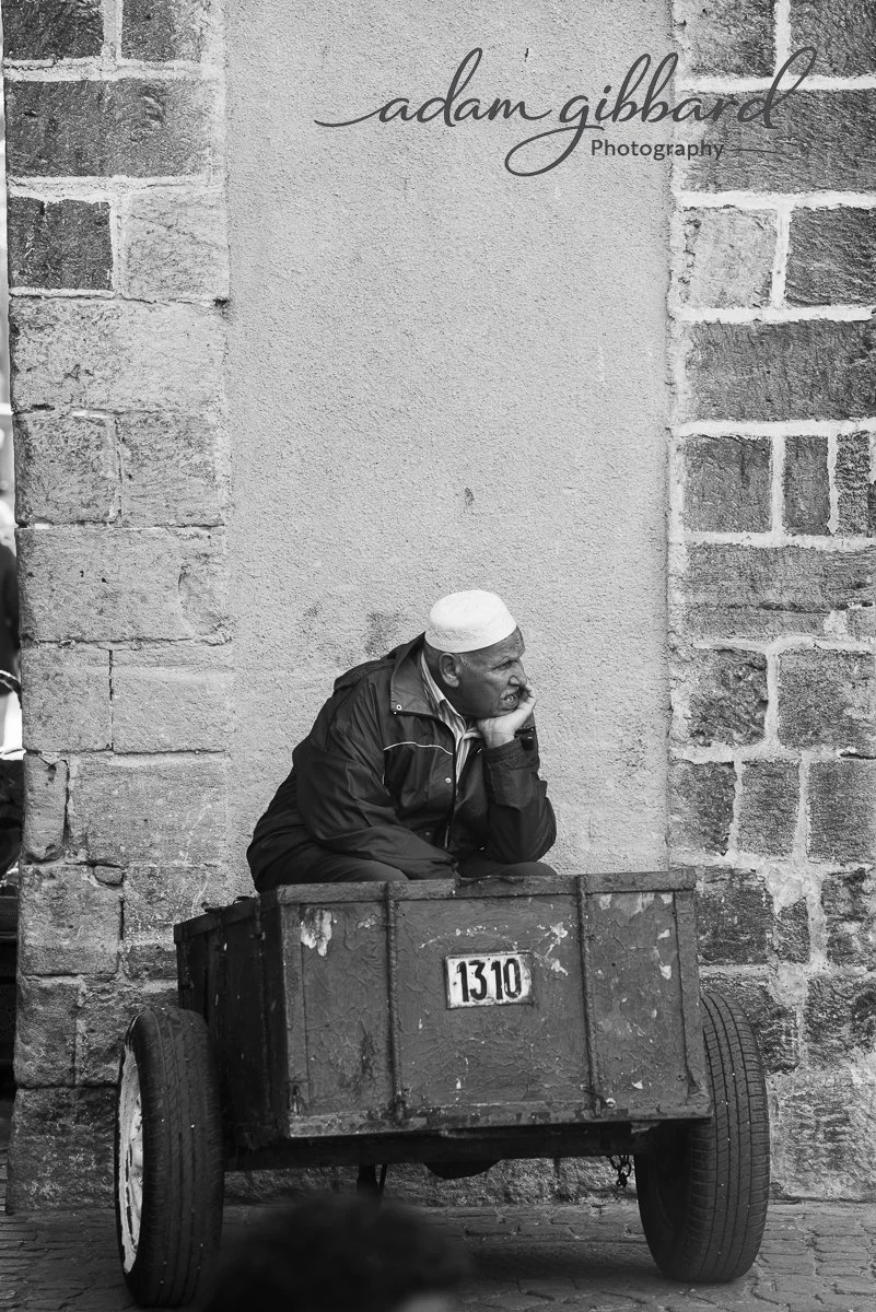 A man sitting in a wooden cart against a stone wall, resting his chin on his hand, wearing a white cap and dark jacket.