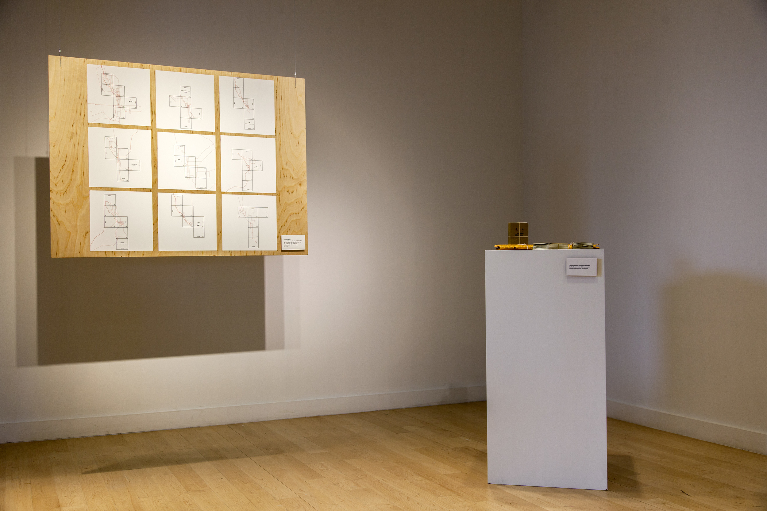 An installation shot of final artwork by Jason Hoylman. Maps representing the paths of his neighbors, and their journals on a pedestal.