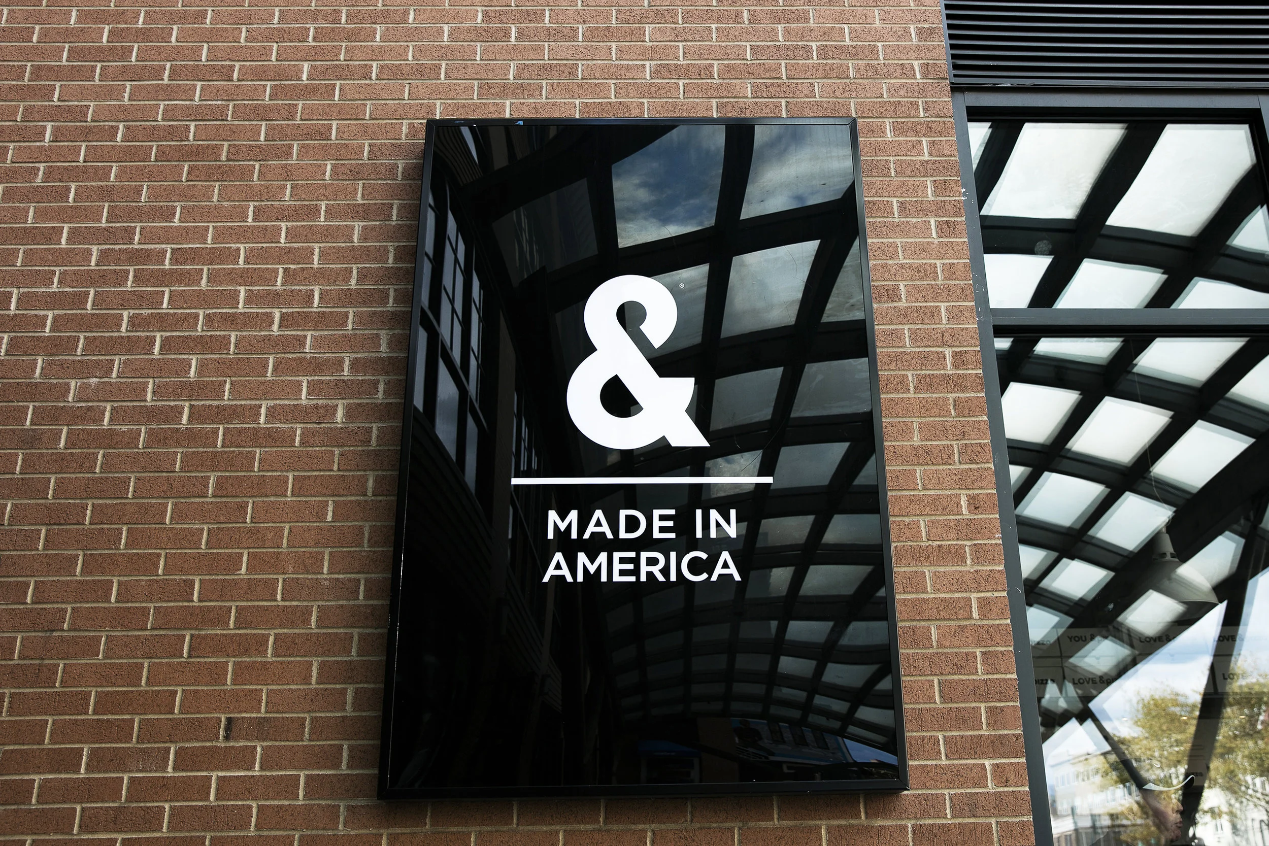 A black sign mounted on a brick wall displays a large white ampersand and the words "MADE IN AMERICA" below it. The sign reflects the glass ceiling and sky above.