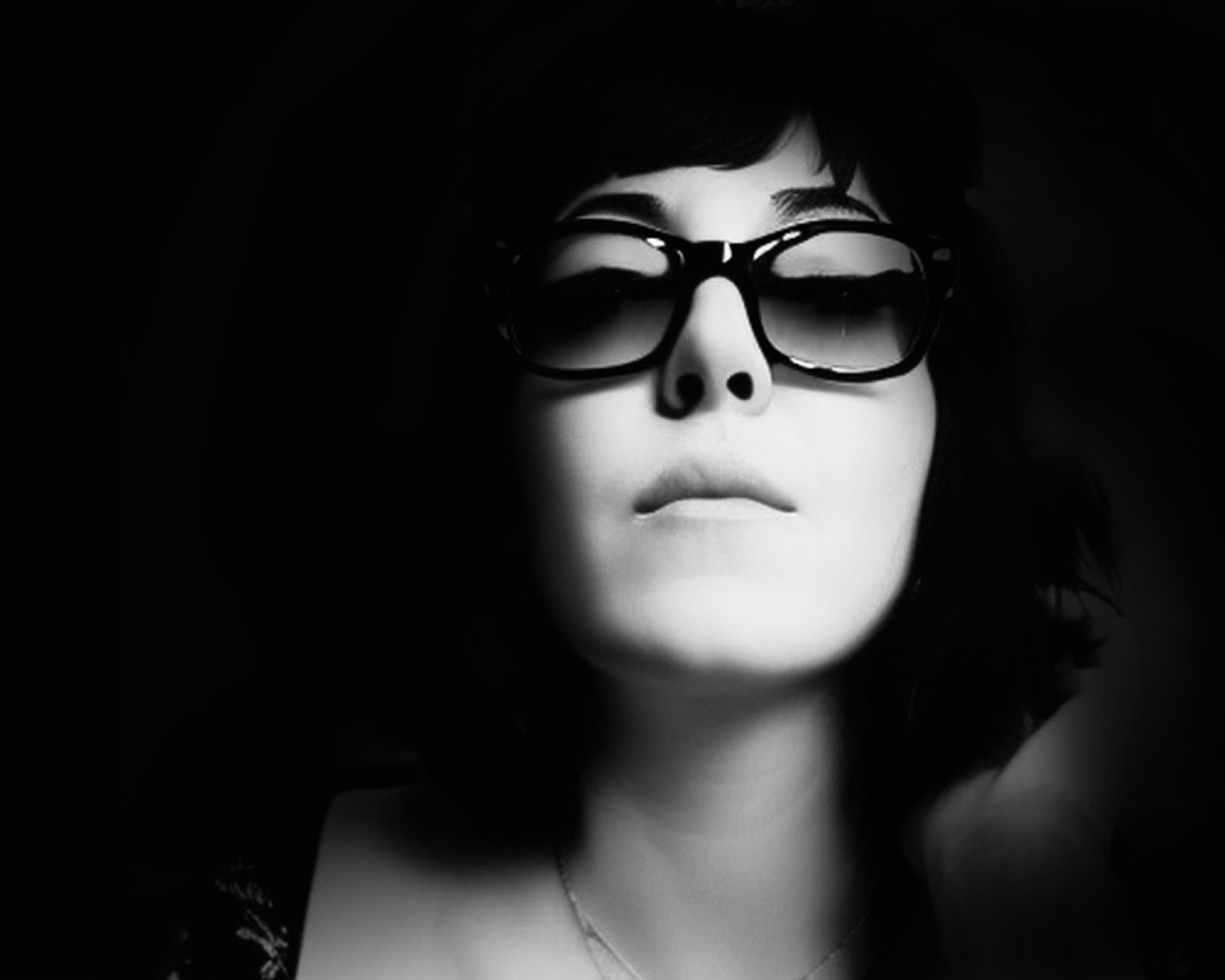Black and white portrait of a woman wearing large sunglasses and a wide-brimmed hat, with dark hair and a necklace, against a dark background.