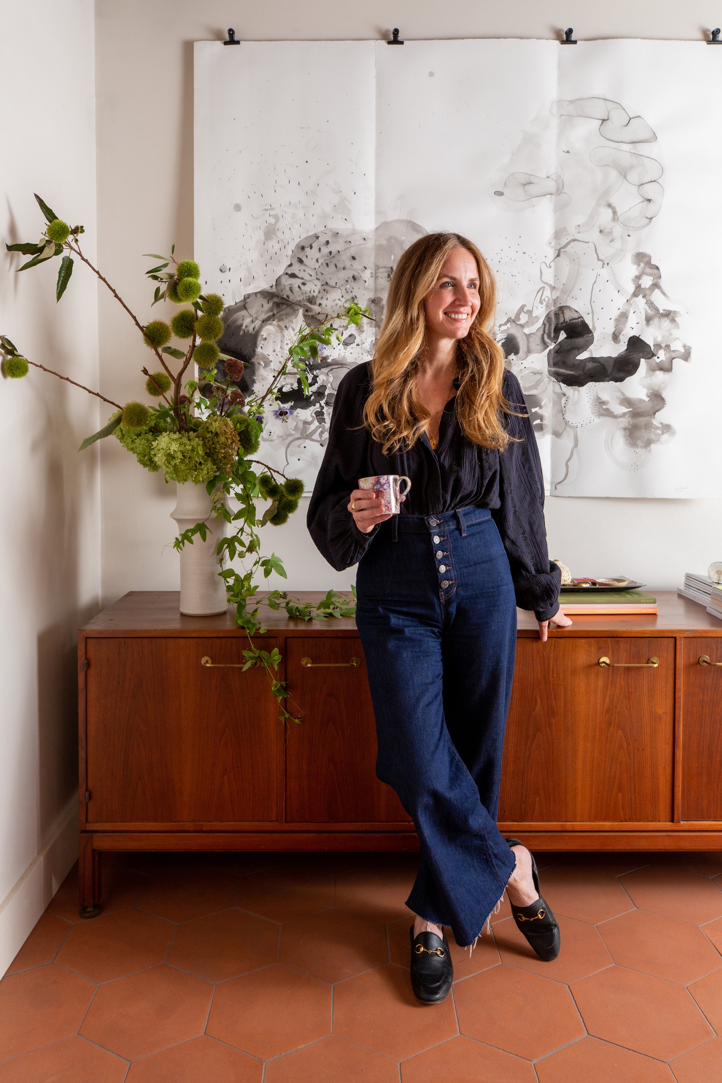 Woman in jeans and blouse smiling and holding coffee mug in stylish interior setting
