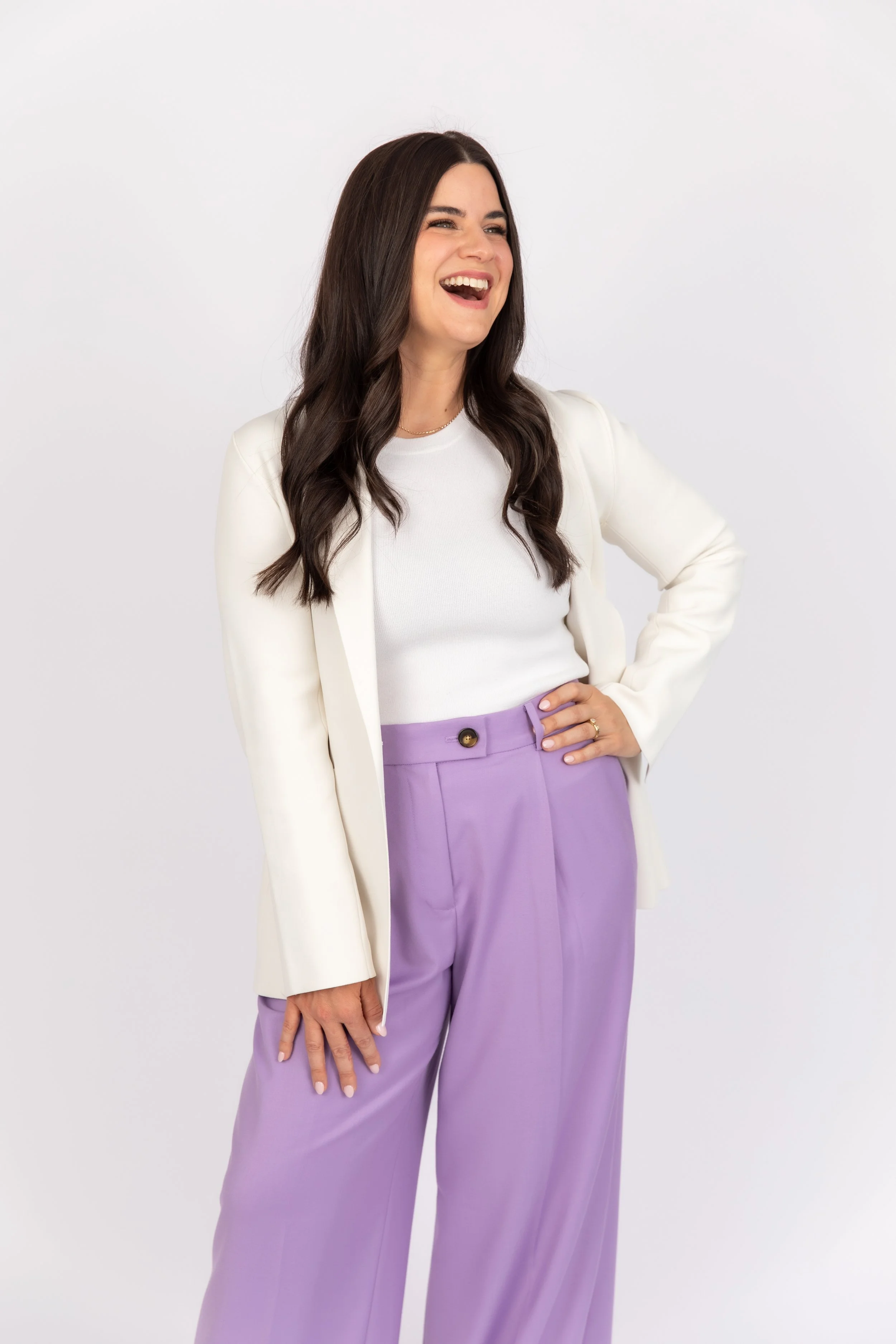 Candid studio headshot of woman in purple pants and white blazer looking to the side with big smile
