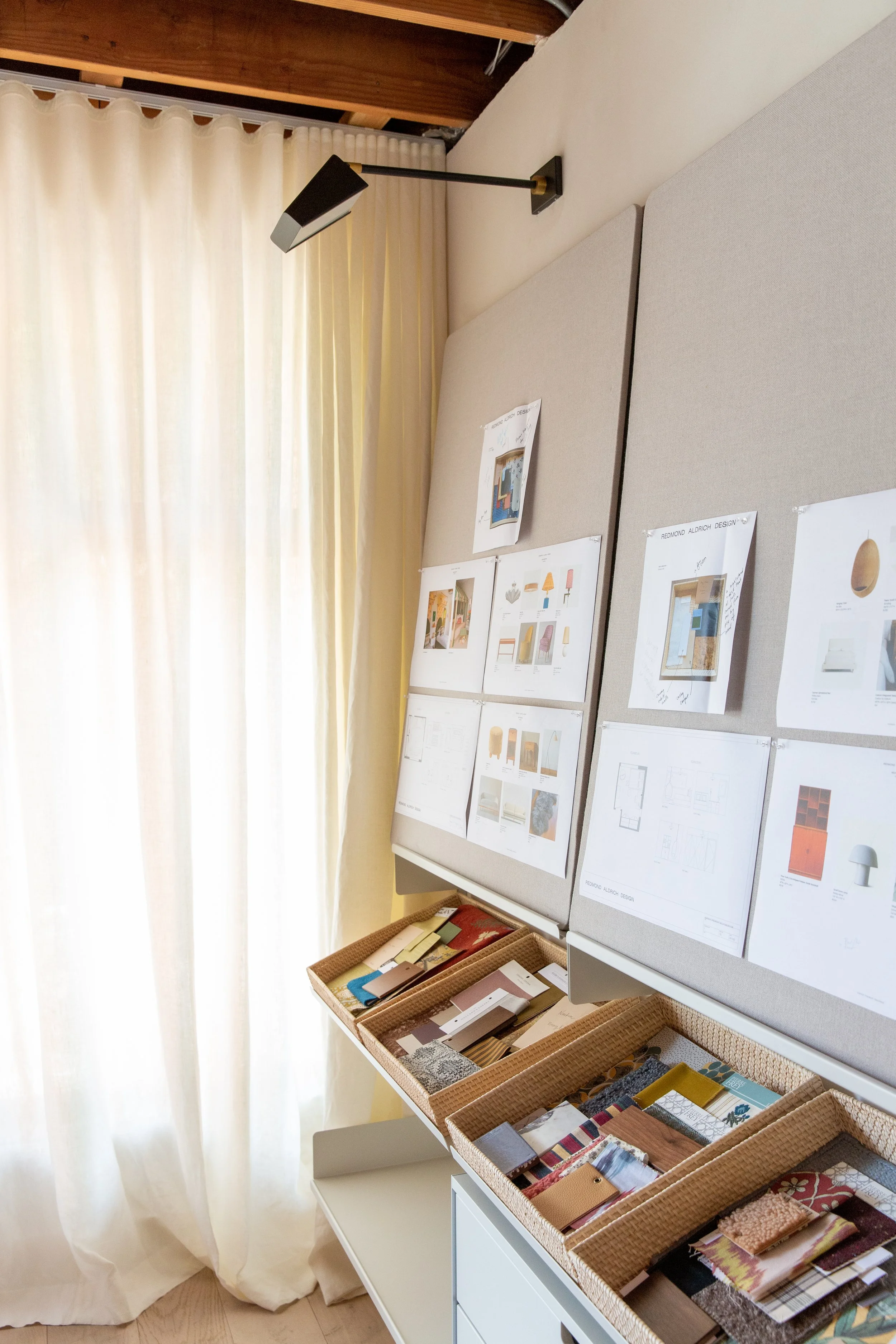 Display of fabric samples and interior design sketches on bulletin boards in a well-lit room with cream curtains.