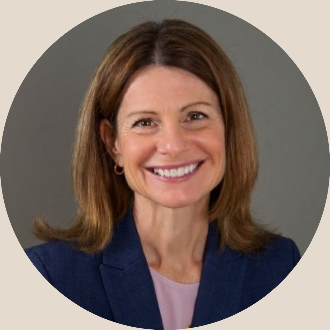 A professional middle aged woman with shoulder-length brown hair smiling in front of a gray background, wearing a navy blazer and light pink top.