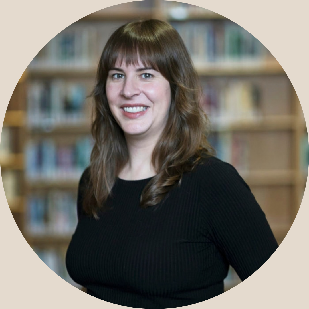 A perimenopausal woman with brown, wavy hair wearing a black top, standing in a library with books on shelves in the background.