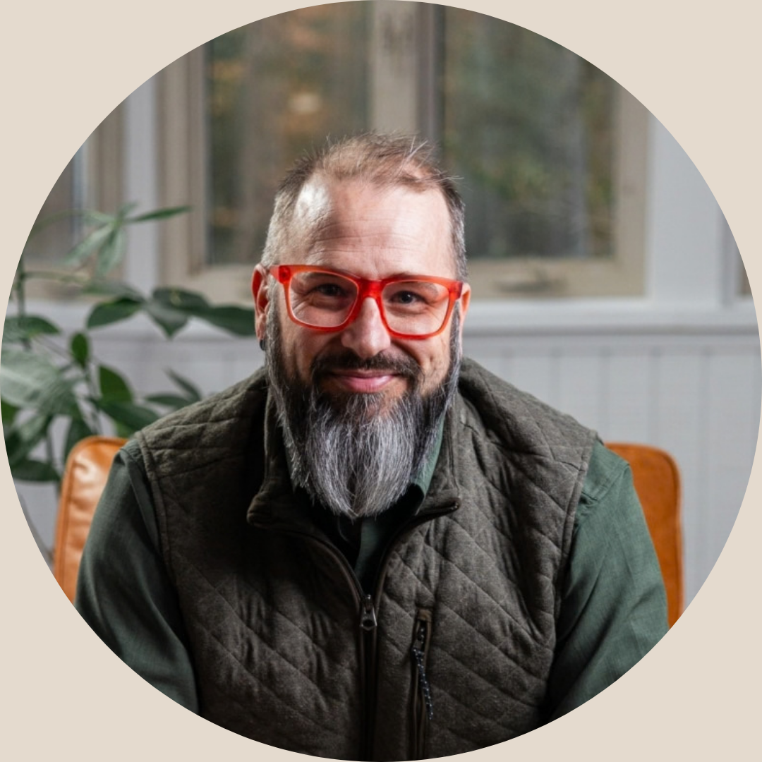 A smiling man with a beard and glasses sitting indoors with a green plant and window in the background.