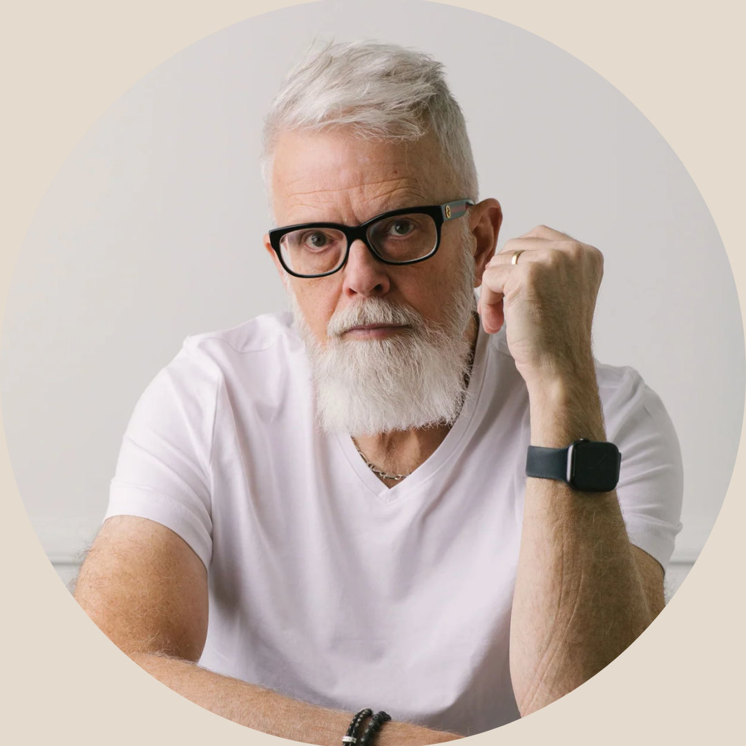 An older man with white hair, glasses, and a beard, wearing a white T-shirt, showing a serious expression with his right fist raised and his left arm resting on a surface.