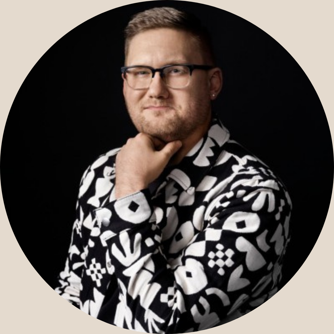Portrait of a man with glasses, short hair, and a beard, wearing a black and white patterned shirt, posing with his hand on his chin against a black background.