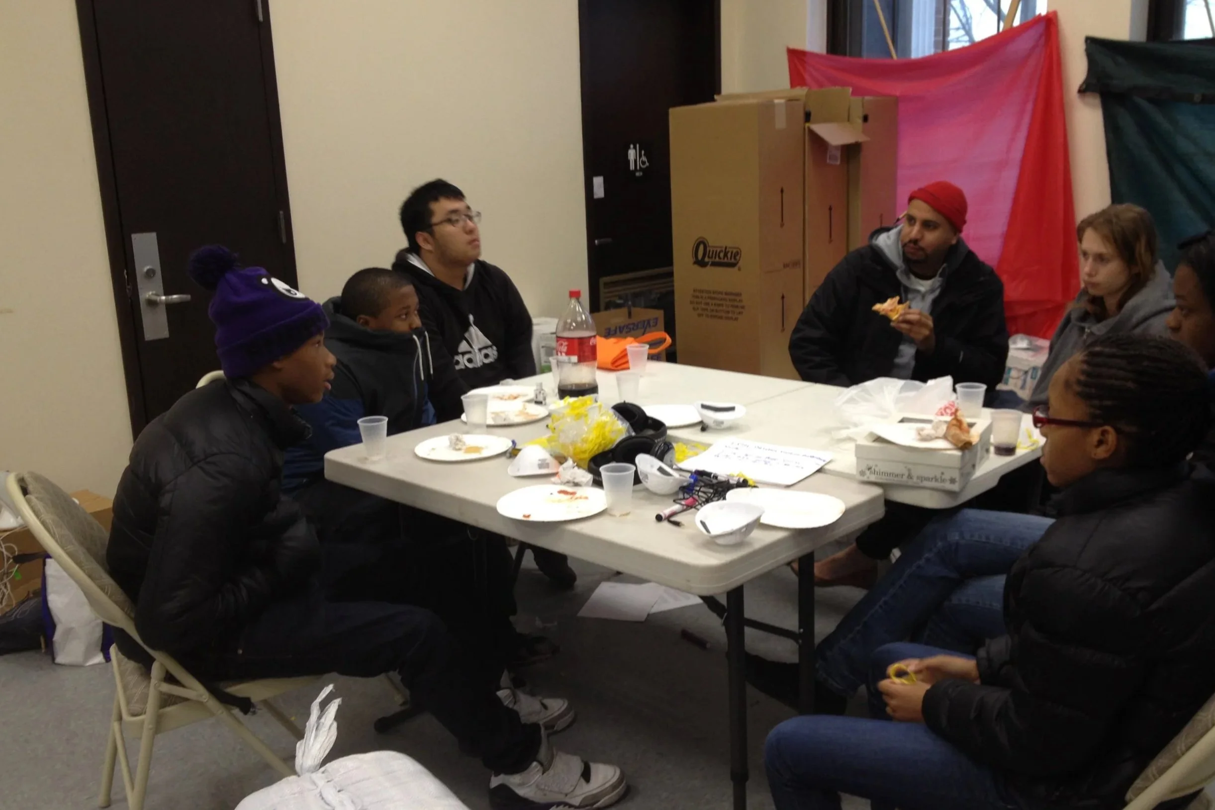 A group of diverse people sitting around a table with food and beverages, in a room decorated with colorful curtains and cardboard boxes.