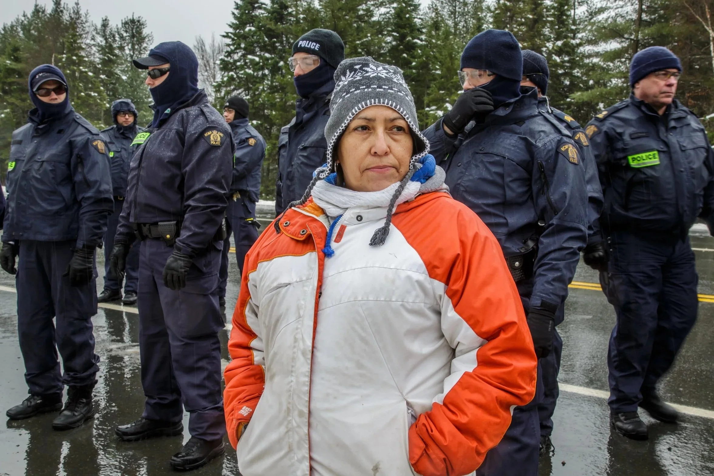 A woman wearing an orange and white jacket with a gray knit hat stands in front of a line of police officers dressed in dark uniforms and wearing protective gear. The scene appears to be outdoors on a rainy day, with trees in the background.