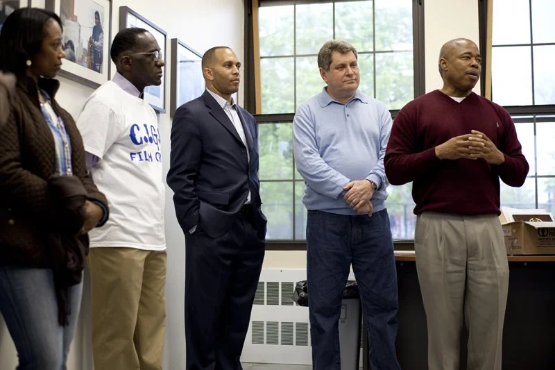 Five people standing indoors in front of large windows, participating in a discussion or presentation, with photo frames on the wall behind them.