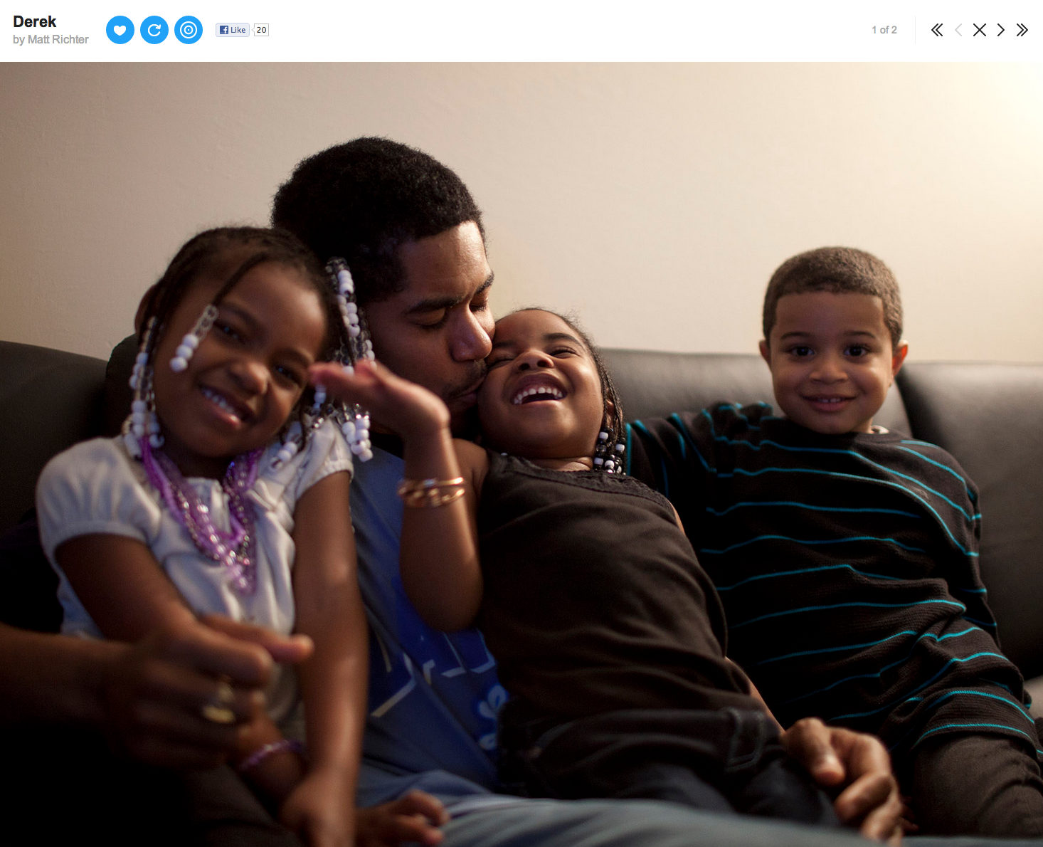 A father and four children sitting on a black sofa, with the father kissing a young girl on the cheek. The children are smiling; the girl with braids and jewelry, the boy on the right with short curly hair and a striped shirt, and two other girls smi