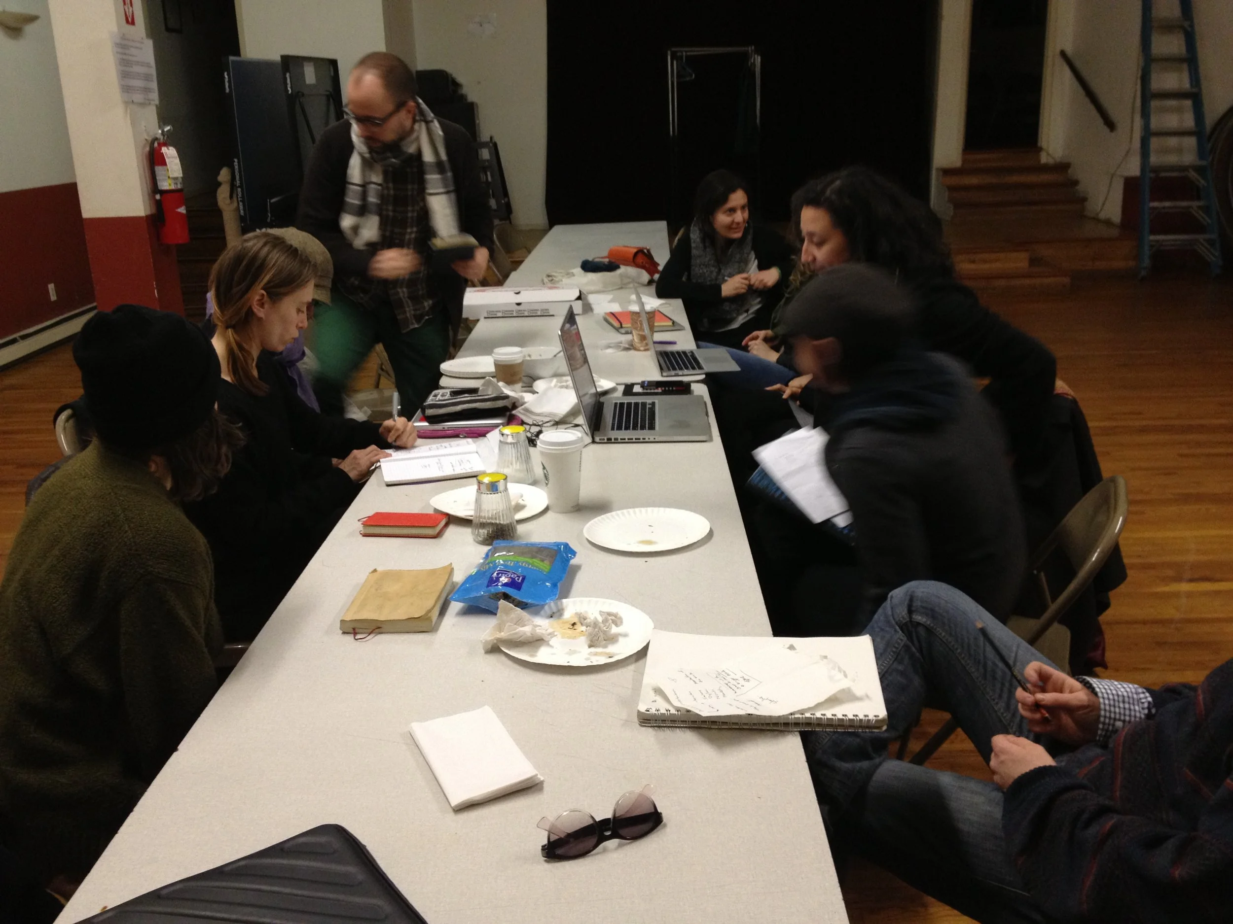 People sitting and standing around a table with laptops, notebooks, food, and coffee cups, in a room with a staircase and a ladder in the background.