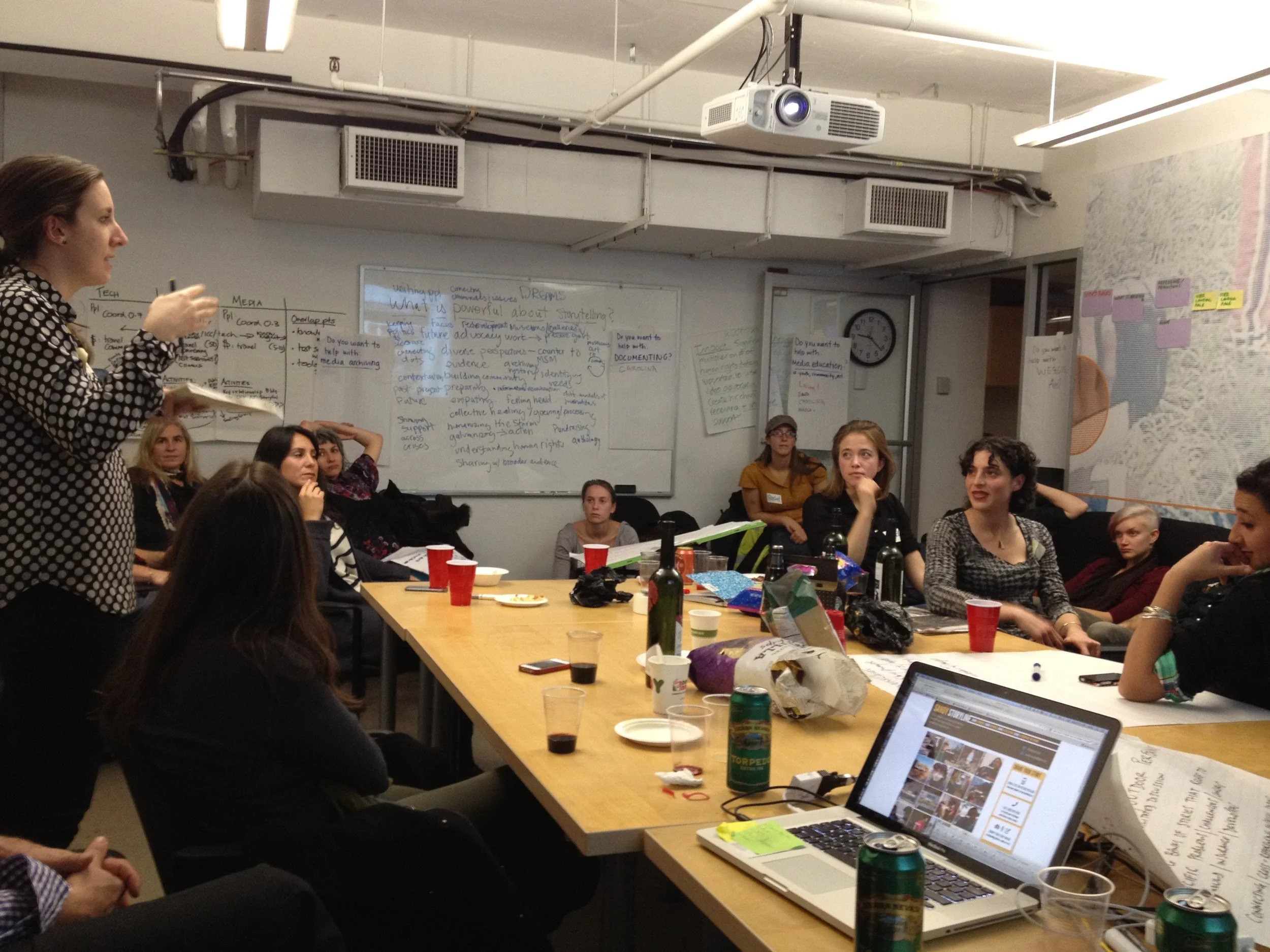 A group of people attending a presentation or workshop in a conference room, with some sitting at tables, and a woman standing and speaking to the group. There are whiteboards with notes, a projector hanging from the ceiling, and various items on the