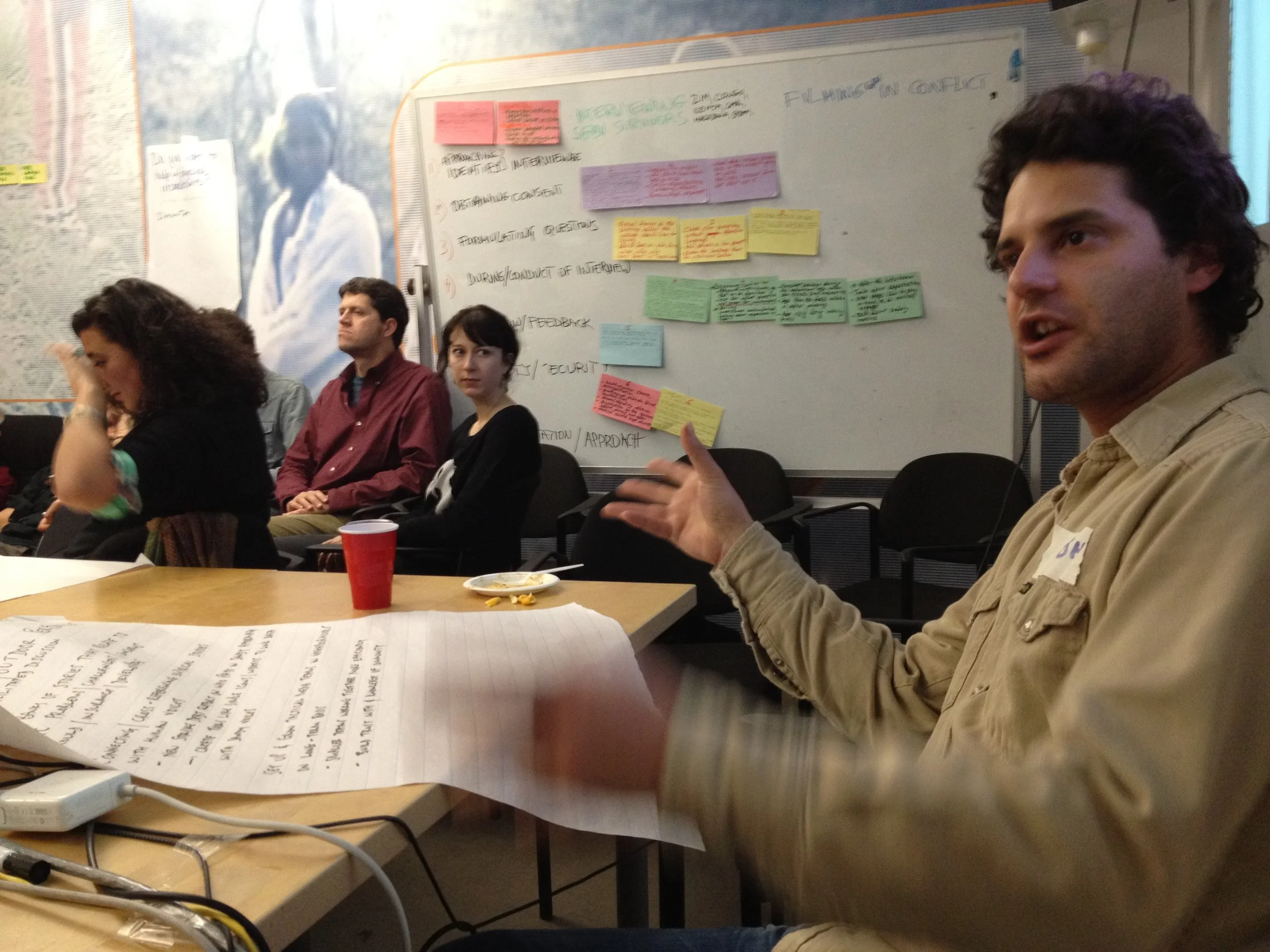 Four people sitting at a table during a meeting or discussion, with a whiteboard behind them filled with handwritten notes and colorful sticky notes, papers, and food items on the table.