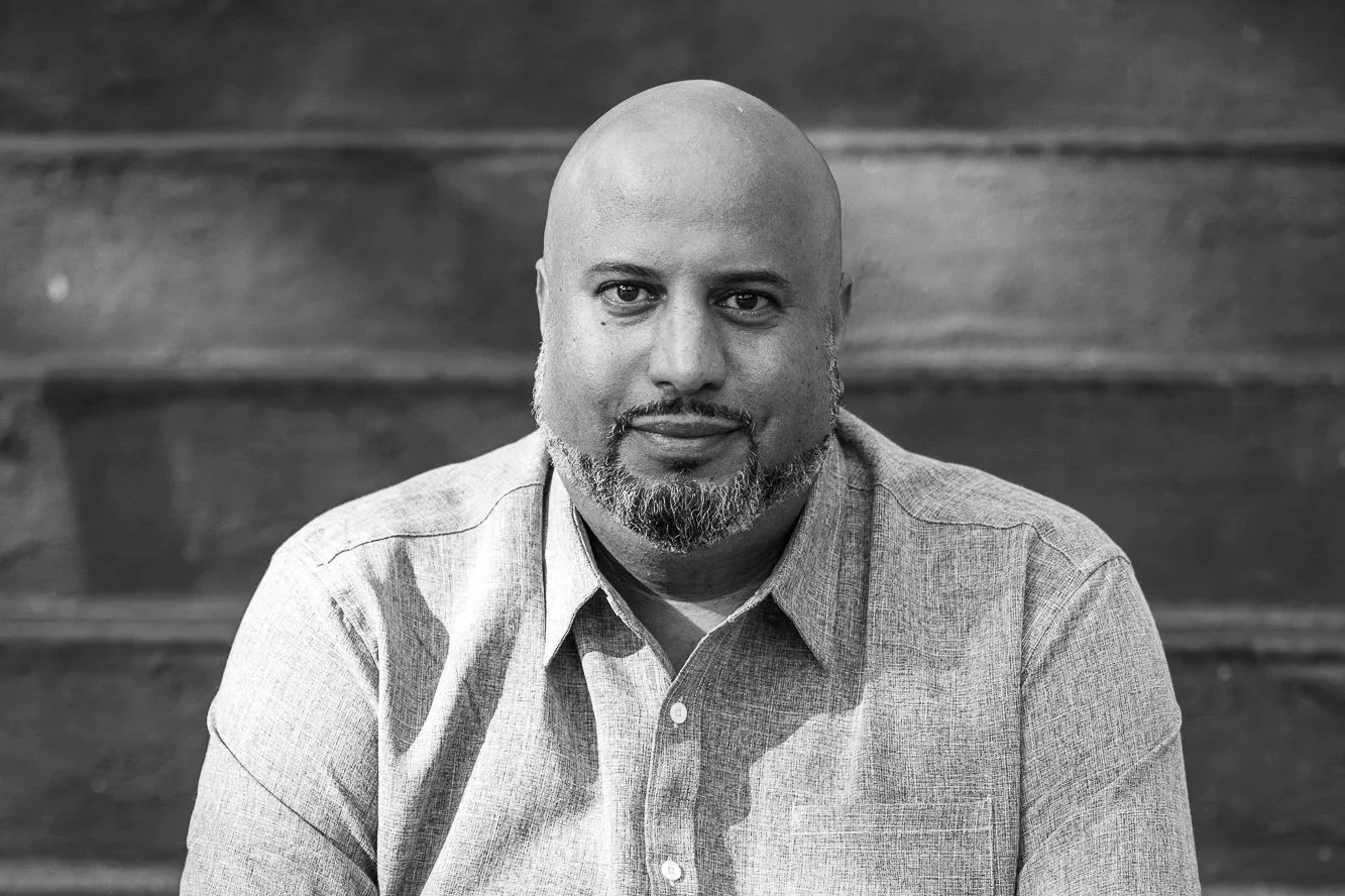 Black and white portrait of a bald man with a beard, wearing a button-up shirt, sitting on wooden stairs.
