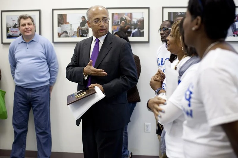 A man in a suit and purple tie is speaking to a group of women in white shirts, inside a room with framed pictures on the wall. Other men are standing nearby.