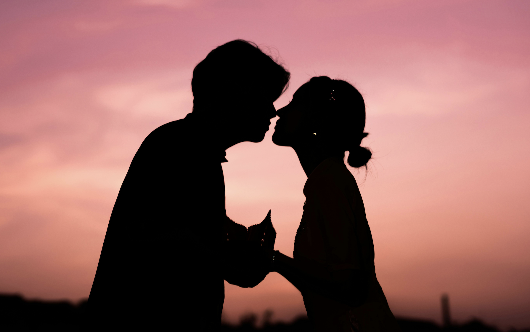Silhouette of two people leaning in for a kiss with a pink and orange sky behind them.