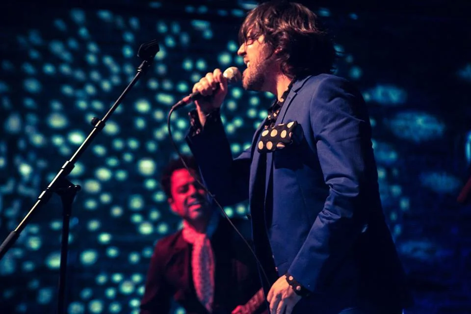 Image Description: Norwood, wearing a blue suit and black and white polka dot tie, sings at a concert. (Photo courtesy of Graham Norwood)
