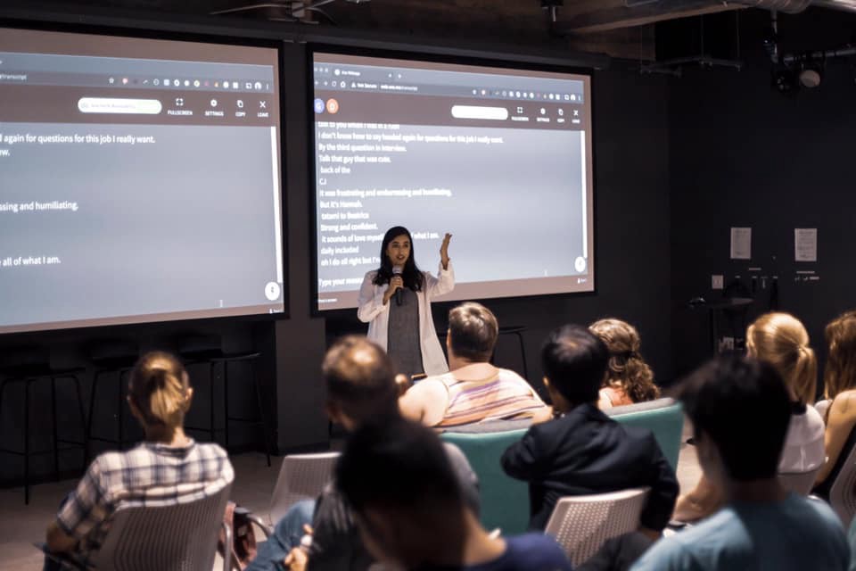 Emon Shakoor speaking into a microphone in front of large screens displaying captions in white. Photo credit: Kai Faust