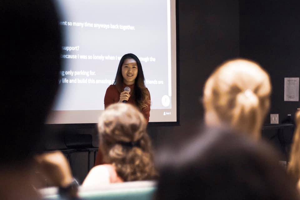 Kwiri Yang speaking into a microphone taken from a seat in the audience and a few out focus heads seated in front of the photographer. Photo credit: Kai Faust