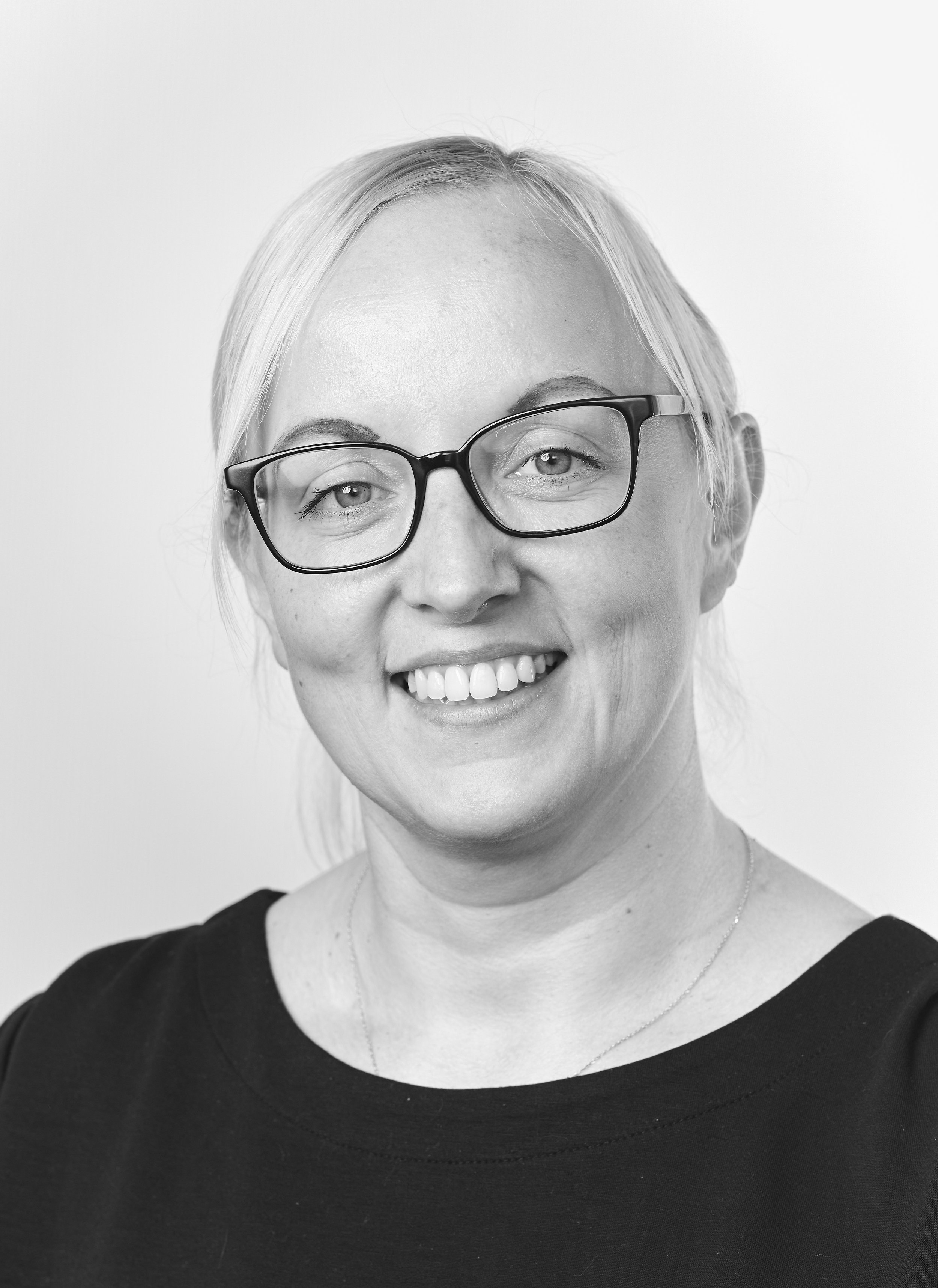 A smiling woman with glasses, blonde hair tied back, wearing a dark top and a delicate necklace.