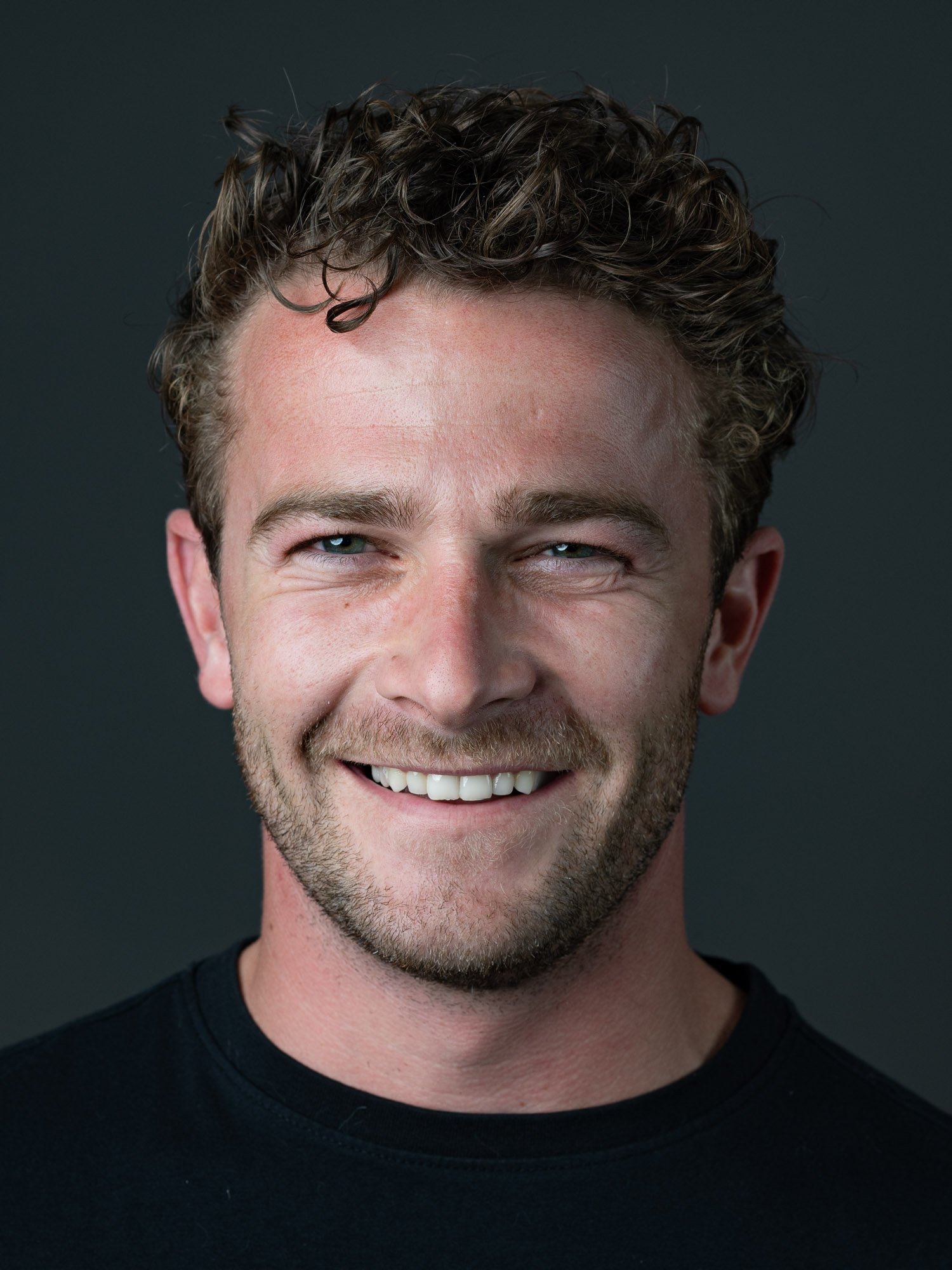Close-up of a smiling man with curly hair, blue eyes, and light skin, wearing a black shirt against a dark background.
